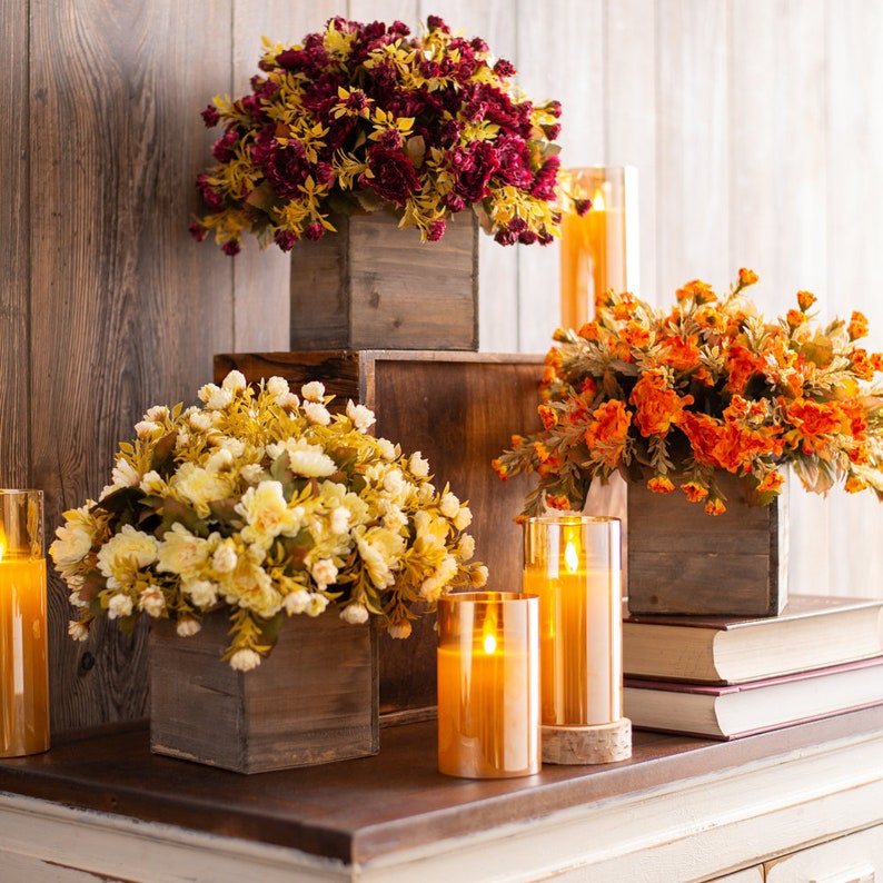 May include: Three wooden boxes with different colored artificial flower arrangements and lit candles. The boxes are stacked on top of each other on a white wooden surface.