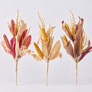 May include: Three decorative floral stems featuring dried corn cobs in shades of red, yellow, and gold, accompanied by dried husks and thin, wispy yellow stalks. Each stem is wrapped at the base, showcasing a natural, harvest-inspired arrangement.
