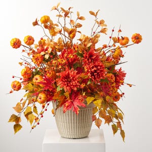 Puede incluir: Arreglo floral otoñal en una maceta clara y texturizada. El arreglo presenta dalias naranjas, pequeñas calabazas y hojas de otoño en tonos rojos, naranjas y amarillos. Las bayas añaden un toque de detalle a la exhibición.