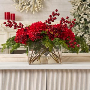 May include: A seasonal floral display in a clear rectangular glass vase. The arrangement includes bright red hydrangeas, berries, and green foliage. The display is set against a neutral background, suggesting a festive or holiday theme.