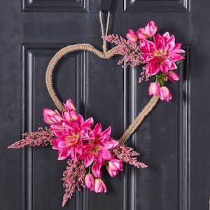 May include: A pink and green floral wreath in the shape of a heart, made with burlap rope and artificial flowers, hangs on a black door.