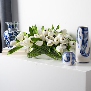 May include: A rectangular glass vase filled with white tulips and green leaves. Two blue and white ceramic vases with floral patterns and a small round vase with a fern design are also present, creating a decorative display.