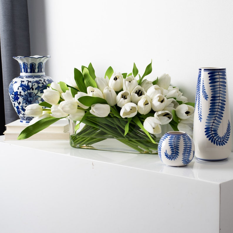May include: A rectangular glass vase filled with white tulips and green leaves. Two blue and white ceramic vases with floral patterns and a small round vase with a fern design are also present, creating a decorative display.