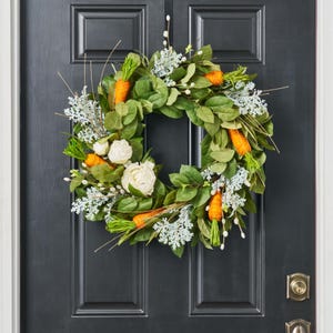 May include: A round Easter wreath with green leaves, white flowers, and orange carrot decorations. The wreath is hanging on a black door. The carrots have green tops and are interspersed throughout the wreath.