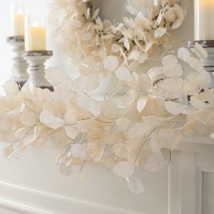 May include: A cream-colored garland of round, papery leaves, draped across a white surface. A matching wreath hangs in the background. Two white candle holders with lit candles are on the left.