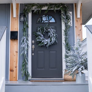 May include: A gray front door with a frosted evergreen wreath and garland. The door is flanked by white railings and a wooden post with a mailbox. The house is painted a dark gray color.