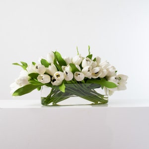 May include: A rectangular glass vase filled with white tulips and green leaves. The arrangement is set against a white background, creating a clean and elegant aesthetic. The tulips are in full bloom.