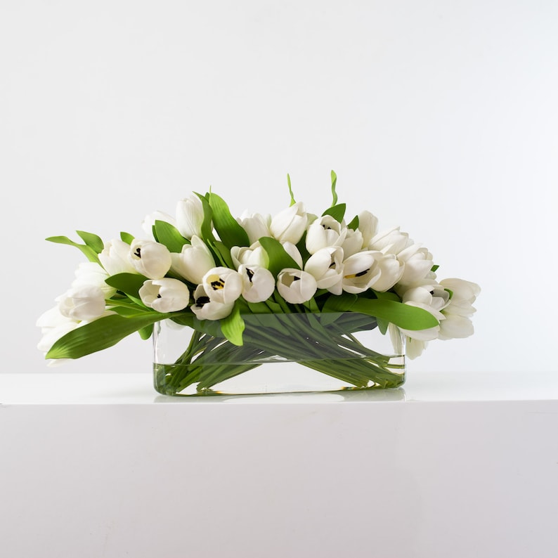 May include: A rectangular glass vase filled with white tulips and green leaves. The arrangement is set against a white background, creating a clean and elegant aesthetic. The tulips are in full bloom.