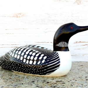 Puede incluir: Figurita de loon de madera tallada a mano con plumas con patrones en blanco y negro y ojos rojos. El loon est&aacute; acostado boca abajo con la cabeza girada hacia un lado.