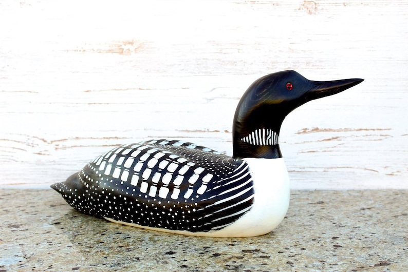 Puede incluir: Figurita de loon de madera tallada a mano con plumas con patrones en blanco y negro y ojos rojos. El loon est&aacute; acostado boca abajo con la cabeza girada hacia un lado.