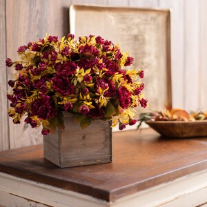 May include: A bouquet of burgundy and yellow flowers in a wooden box. The flowers are arranged in a rustic style and are sitting on a wooden table.