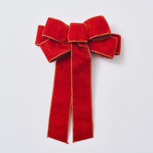 Puede incluir: Un lazo grande de terciopelo rojo con ribete dorado. El lazo tiene múltiples bucles y dos largas colas rectangulares. El lazo probablemente se usa para decoración, como en un regalo o como decoración navideña.