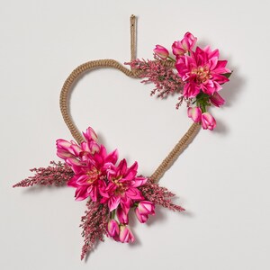 May include: A heart-shaped wreath made of natural twine is decorated with pink flowers. The wreath is perfect for a spring or summer door decoration.