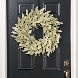 May include: A circular wreath composed of numerous pale green, pointed leaves arranged in a dense, overlapping pattern. The foliage has a muted, matte finish, creating a full and rounded appearance that forms a consistent ring shape throughout.
