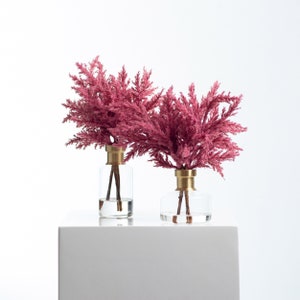 以下が含まれることがあります： 金色の蓋が付いた透明なガラスの花瓶2つに、ピンクのドライフラワーが活けられています。花瓶は白い台の上に置かれています。
