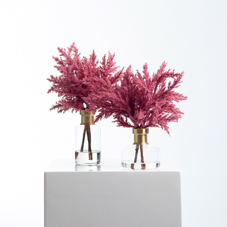以下が含まれることがあります： 金色の蓋が付いた透明なガラスの花瓶2つに、ピンクのドライフラワーが活けられています。花瓶は白い台の上に置かれています。