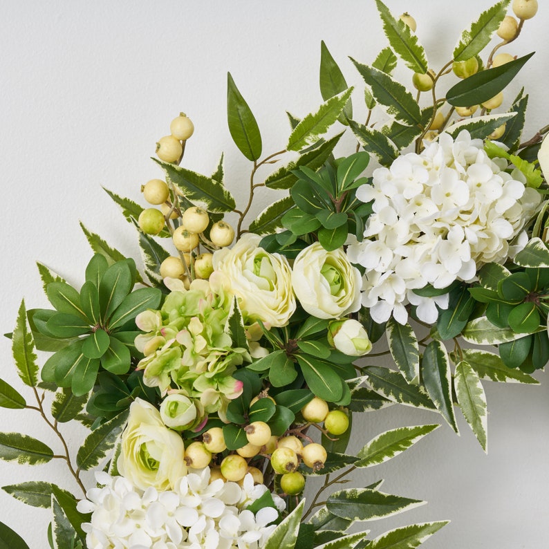 Peut inclure: Une couronne florale verte et blanche avec des hortensias blancs, des renoncules cr&egrave;me et des baies vertes.