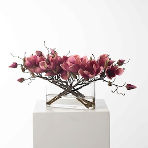 May include: A bouquet of pink magnolia flowers arranged in a clear glass vase. The flowers are on a white pedestal.
