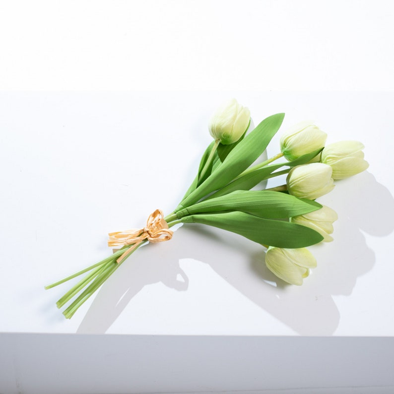 以下が含まれることがあります： 金色のリボンで結ばれた、緑色の茎を持つ人工の白いチューリップのブーケです。