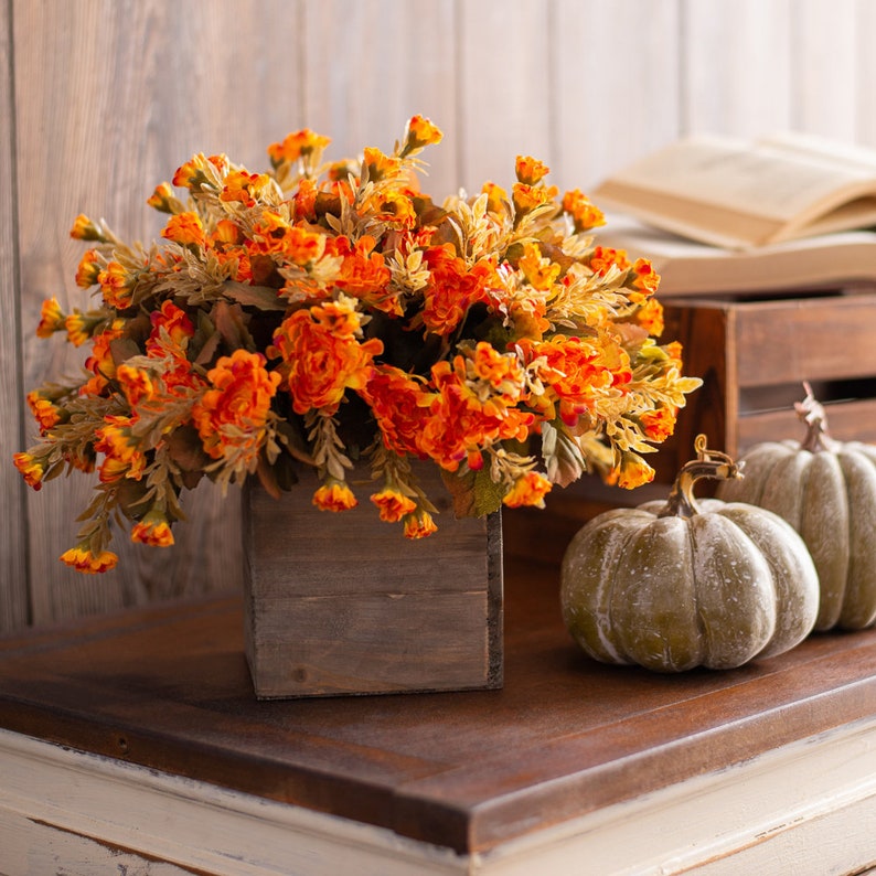 May include: A wooden box planter filled with a bouquet of orange and yellow silk flowers. Two green pumpkins sit on a wooden table in front of the planter.
