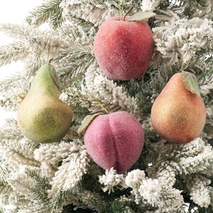 Peut inclure: Quatre ornements en forme de fruits suspendus à une branche de sapin floquée. L'ensemble comprend une pomme rouge, une poire verte, une poire bronze et une pêche rose, toutes avec une finition texturée et pailletée et de petites feuilles vertes.