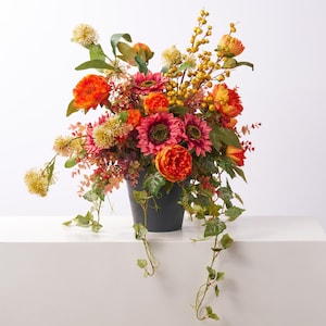 May include: A floral arrangement in a dark gray pot featuring orange and deep pink sunflowers, vibrant orange peonies, yellow berry sprigs, and trailing green ivy leaves. The composition includes various textured filler flowers and foliage.