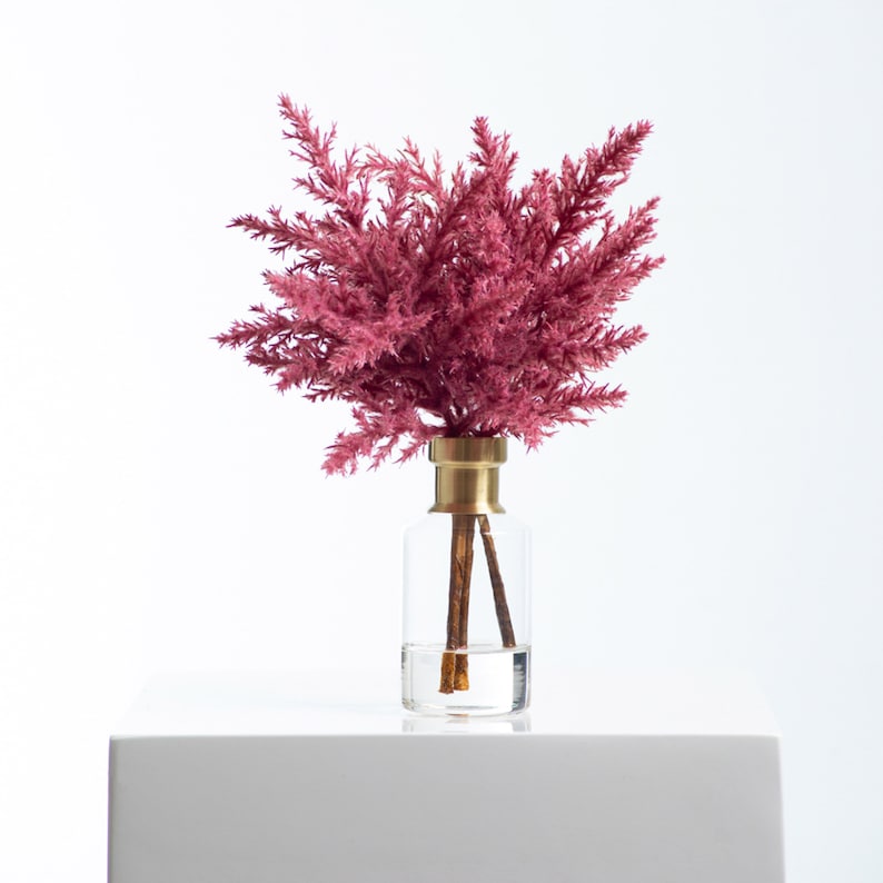 以下が含まれることがあります： 金色の蓋が付いた透明なガラスの花瓶に、ピンクのドライフラワーのフラワーアレンジメントが飾られています。花瓶は白い表面の上に置かれています。