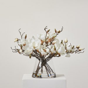 May include: A bouquet of white magnolia flowers arranged in a clear glass vase with a textured surface. The vase is sitting on a white surface.