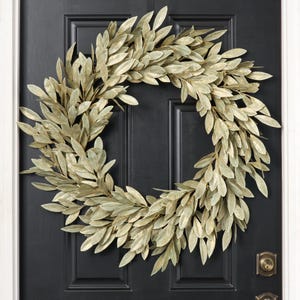 May include: A circular wreath composed of numerous overlapping pale gold, metallic-finished leaves arranged in a dense, layered pattern. The foliage features pointed, elongated shapes with a subtle sheen, creating a textured, full appearance.