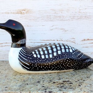 Puede incluir: Figurita de loon de madera tallada a mano con marcas pintadas en blanco y negro. El loon tiene ojos rojos y un pico largo y delgado.