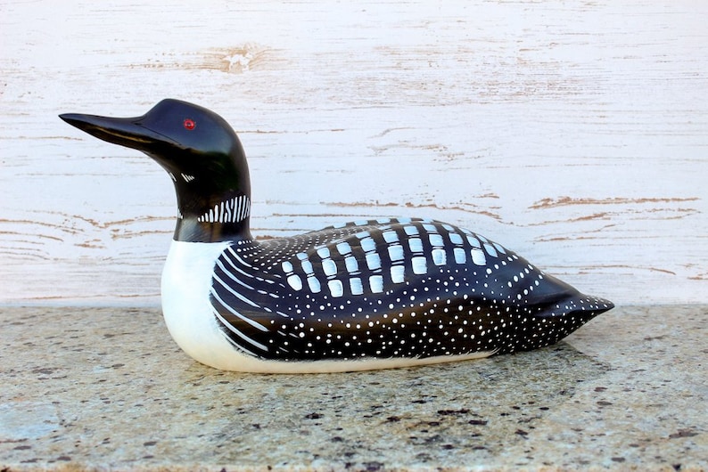 Puede incluir: Figurita de loon de madera tallada a mano con marcas pintadas en blanco y negro. El loon tiene ojos rojos y un pico largo y delgado.