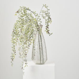 May include: A clear glass vase with a textured, mercury-like finish holds cascading greenery with small, reddish-purple accents. The vase has a teardrop shape and sits on a white surface, creating a minimalist aesthetic.