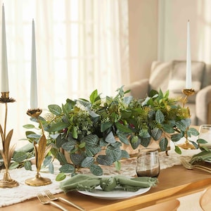 Puede incluir: Un centro de mesa de madera lleno de plantas verdes de hoja y bayas. El centro de mesa está sobre una mesa de madera con candelabros blancos y servilletas verdes.