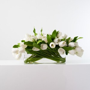 May include: A rectangular glass vase filled with white tulips and green leaves. The flowers are arranged in a dense cluster, creating a full and vibrant display. The vase sits on a white surface against a white background.