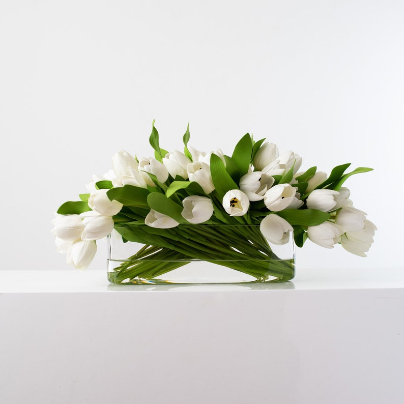 May include: A rectangular glass vase filled with white tulips and green leaves. The flowers are arranged in a dense cluster, creating a full and vibrant display. The vase sits on a white surface against a white background.