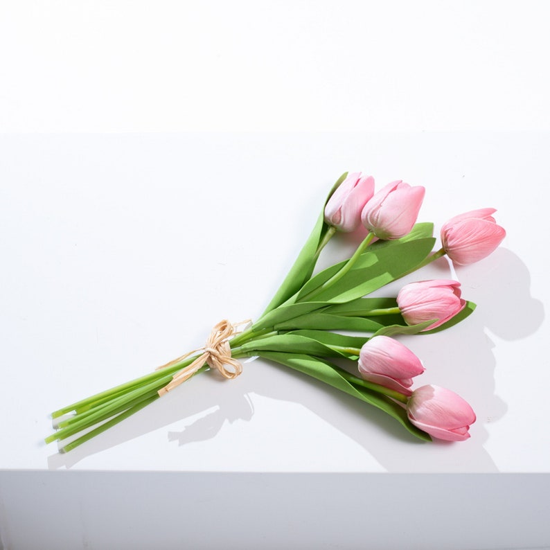 以下が含まれることがあります： 茶色のリボンで結ばれた、緑色の茎と葉を持つピンクのチューリップのブーケです。