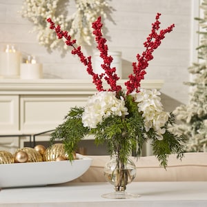 May include: A clear glass vase holds a floral arrangement. The arrangement features white hydrangeas, green sprigs, and red berry stems. The vase sits on a white surface, with gold ornaments in a white bowl in the background.