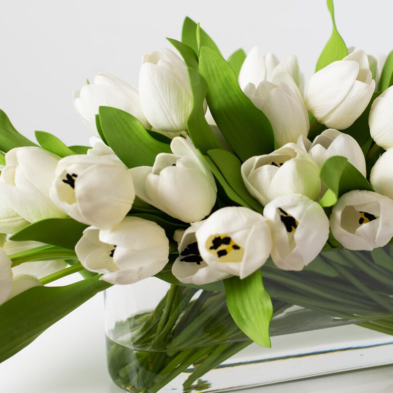 May include: A bouquet of white tulips with green leaves in a clear glass vase. The tulips are tightly closed, with black and yellow centers visible in some blooms. The arrangement is set against a white background.