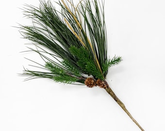 May include: A sprig of artificial greenery featuring long, dark green pine needles, small evergreen branches, and two small pine cones. The stem is a light brown color. The sprig is set against a white background.