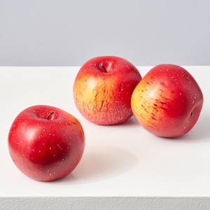 May include: Three artificial apples with a glossy red finish and yellow-orange streaks. The apples have small white speckles and are arranged on a white surface. The stems are brown. These decorative apples are a fall-themed home decor item.