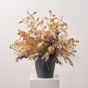 May include: A decorative arrangement of metallic gold and bronze-toned faux foliage, berries, and two spherical woven gold ornaments. The botanical display is contained within a matte black tapered pot, creating a dense, textured centerpiece.