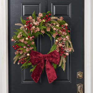 May include: A circular wreath made of green evergreen branches, decorated with metallic rose gold flowers, shiny red berries, and small gold and dark red ornaments. A large, deep red velvet bow is attached to the bottom of the wreath.
