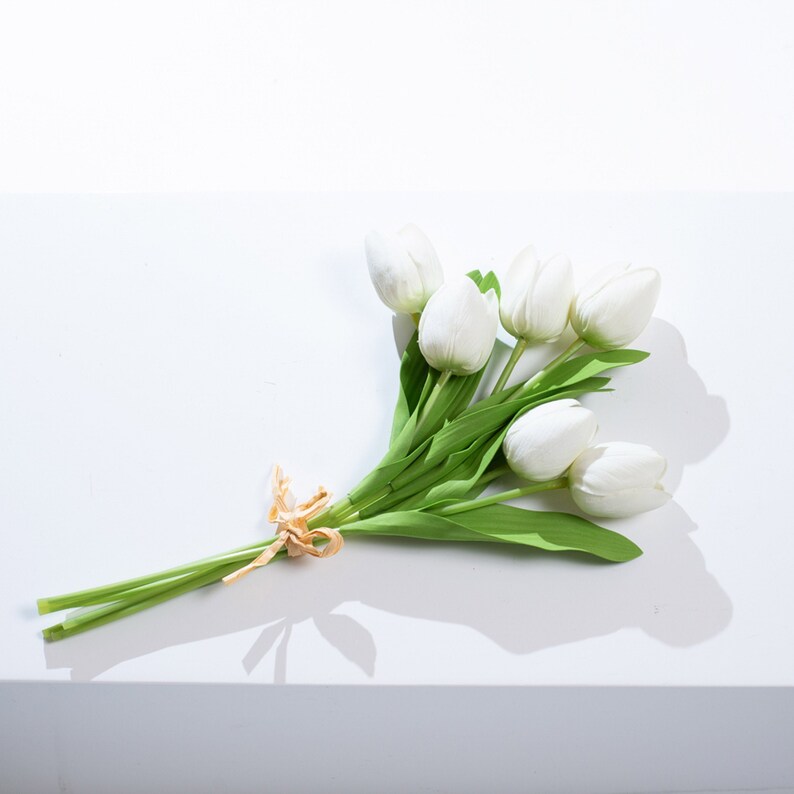 以下が含まれることがあります： 茶色のリボンで結ばれた白い造花チューリップのブーケ。チューリップは緩やかな自然なスタイルでアレンジされています。