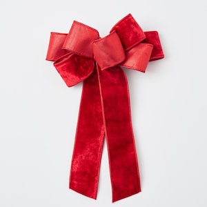 Puede incluir: Un gran lazo festivo de terciopelo rojo con largas colas fluidas. El lazo presenta múltiples bucles y una apariencia rica y texturizada, perfecto para decoraciones navideñas o envoltorios de regalos. El lazo mide aproximadamente 30 cm de ancho.