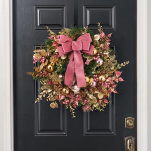 May include: A decorative wreath featuring green pine needles, gold-painted leaves, pinecones, and shiny gold and pink ornaments. A large, dusty rose velvet bow is centred at the top, with long tails hanging down over the foliage.