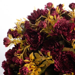 May include: A bouquet of burgundy silk flowers with yellow foliage. The flowers are arranged in a circular shape and have a velvety texture.