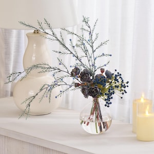 May include: A clear glass spherical vase containing a floral arrangement of dark blue and grey-toned flowers, small berries, and thin, wispy green branches. The stems are visible through the clear glass, which is filled with a small amount of water.