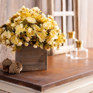 May include: A wooden box filled with a bouquet of cream-colored artificial flowers. The flowers are arranged in a square wooden box with a rustic finish. The box is sitting on a wooden table.