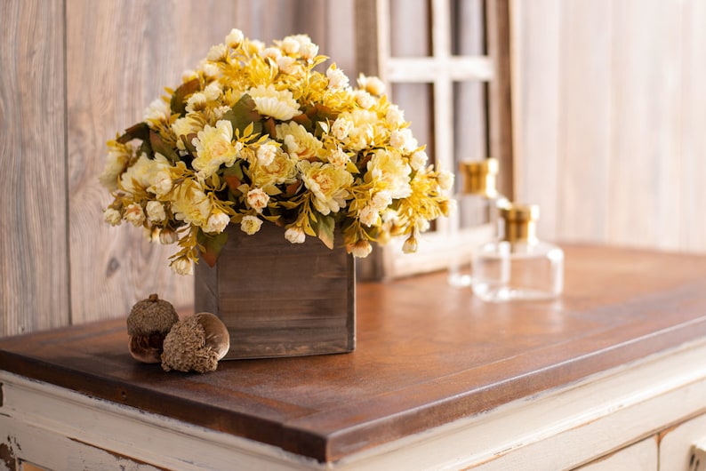 May include: A wooden box filled with a bouquet of cream-colored artificial flowers. The flowers are arranged in a square wooden box with a rustic finish. The box is sitting on a wooden table.