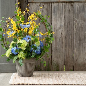 May include: A gray planter with a circular metal frame filled with a floral arrangement of blue and yellow flowers. The flowers are arranged in a circle around the planter, and the metal frame is visible behind the flowers.